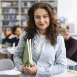 Szkolenia i webinary z kadr i płac w oświacie – kobieta z książkami w bibliotece, w tle dorośli przy stołach